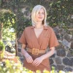 Blonde wig styled on a woman outdoors with greenery and stone wall background.